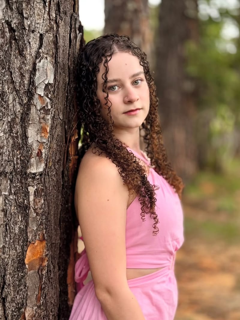 Jovem de vestido rosa encostada em uma árvore, olhando para a câmera